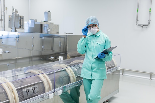 Caucasian factory worker in blue lab suit with tablet in hands stand near manufacture machine hand on glasses