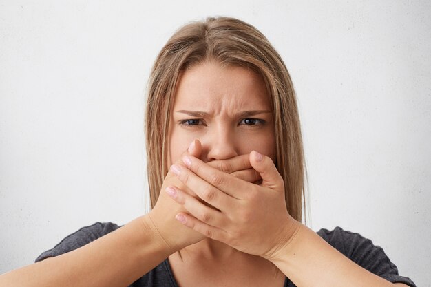 Caucasian fair-haired woman having angry look frowning her eyebrows covering mouth with hands trying to hold tongue. Stylish woman trying to keep silence and not telling secret