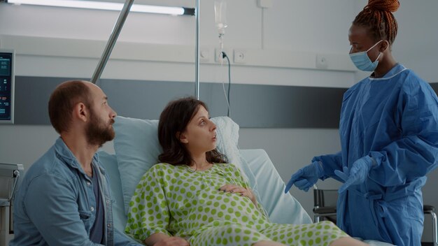産科病棟で赤ちゃんを期待している白人家族。ベッドに座って出産と援助についてアフリカ系アメリカ人の看護師と話している若い妊婦。出産を待っている患者