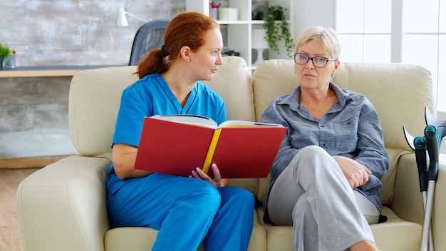 Caucasian female assistant in nursing home reading a book to an elderly retired woman who sits on the couch