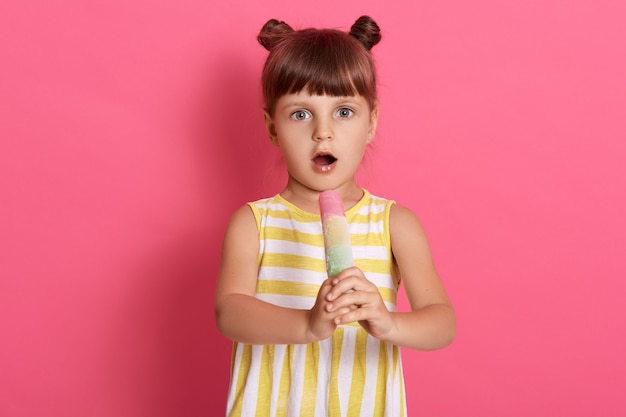 Caucasian female child posing with widely opened mouth, wearing striped summer dress, being shocked, looks surprised, posing with big eyes against pink wall.