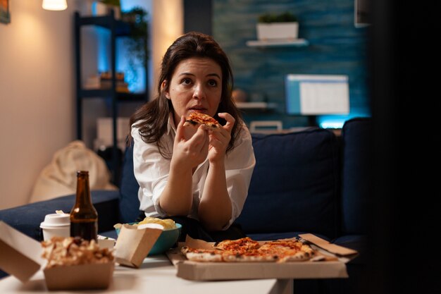 Кавказская женщина держит вкусный кусок пиццы и ест еду на вынос во время просмотра комедии