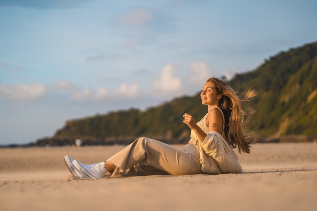 해변에 앉아 있는 아름다운 일몰에 양모 작물을 입은 백인 여성