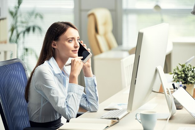 Caucasian female office worker sitting at desk in front of screen and talking on smartphone