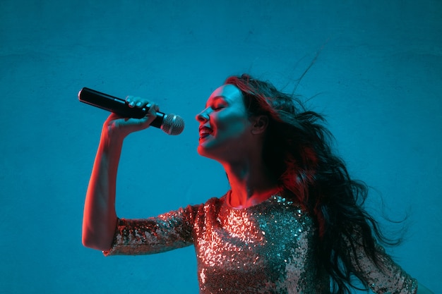 Портрет кавказской певицы, изолированные на синем фоне студии в неоновом свете. Красивая женская модель в ярком платье с микрофоном. Понятие человеческих эмоций, выражение лица, реклама, музыка, искусство.