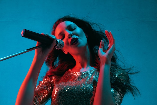 Ritratto di cantante femminile caucasico isolato su sfondo blu studio in luce al neon. bellissimo modello femminile in abito luminoso con microfono. concetto di emozioni umane, espressione facciale, annuncio, musica, arte.