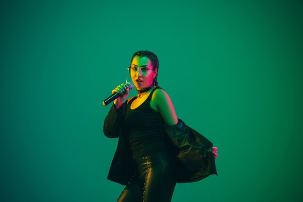Caucasian female singer portrait isolated on green  wall in neon light. Beautiful female model in black wear with microphone. Concept of human emotions, facial expression, ad, music, art.