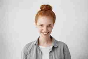 Free photo caucasian female with ginger hair bun looking with her blue charming eyes demonstrating her sincere smile. attractive natural redhead female being glad