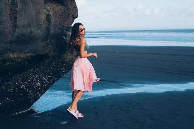 Caucasian fit tanned woman in tiny top and pink skirt on black sand beach