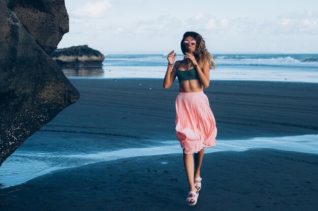 Caucasian fit tanned woman in tiny top and pink skirt on black sand beach