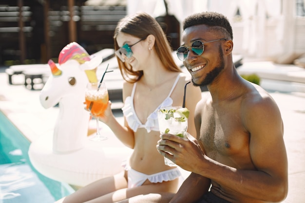 Бесплатное фото Кавказская девушка и ее афро-американский парень отдыхают у бассейна. девушка в белых купальниках и солнцезащитных очках