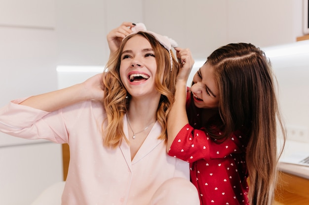 Caucasian glad girl in pink eyemask laughing while posing in kitchen. Indoor photo of pretty sisters joking in morning.