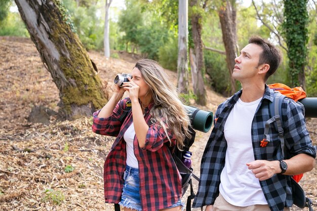Caucasian hikers taking photo, walking or trekking on forest path surrounded with trees. Pretty woman holding camera, shooting and hiking with handsome man. Tourism, adventure and vacation concept