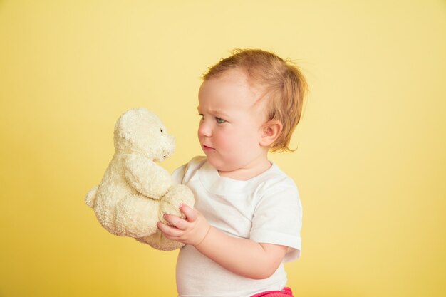 Caucasian little girl, children isolated on yellow studio background. Portrait of cute and adorable kid, baby playing with teddy bear. Concept of childhood, family, happiness, new life, education.