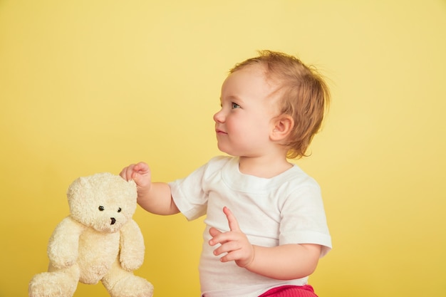 Caucasian little girl, children isolated on yellow studio background. Portrait of cute and adorable kid, baby playing with teddy bear. Concept of childhood, family, happiness, new life, education.