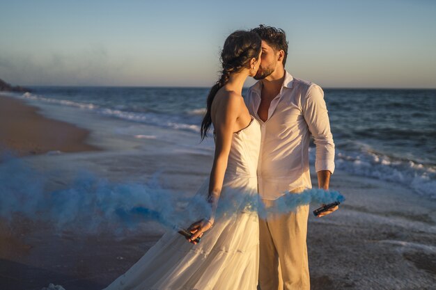 Caucasian loving couple holding a blue colored smoke and kissing in the beach during a wedding