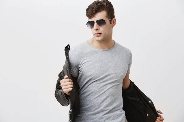 Caucasian male model with appeal look posing indoors . Stylish handsome attractive man with trendy haircut dressed in black leather jacket, wearing sunglasses.