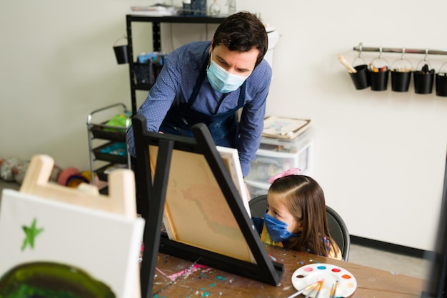 Caucasian male teacher is instructing a girl child how to paint on a canvas for her class in art school for kids