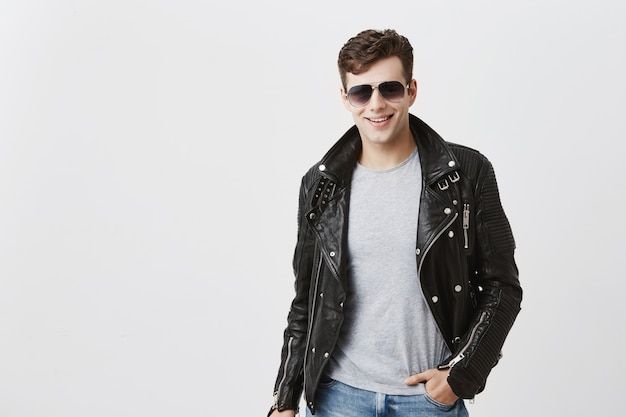 Caucasian male with appeal look, smiling broadly with white even teeth, posing indoors. Stylish handsome attractive man with trendy haircut dressed in black leather jacket,with sunglasses on.