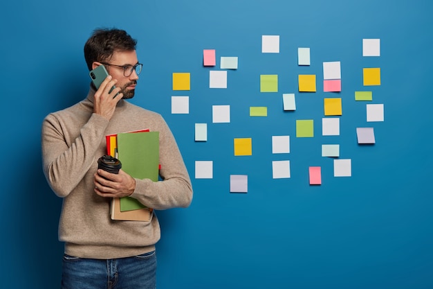 Free photo caucasian man has creative approach to organizing work, leaves colorful stickers on wall, discusses working schedule with partner via smartphone