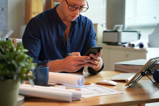 Caucasian man sitting at desk with rolled-up papers and plans and using smartphone