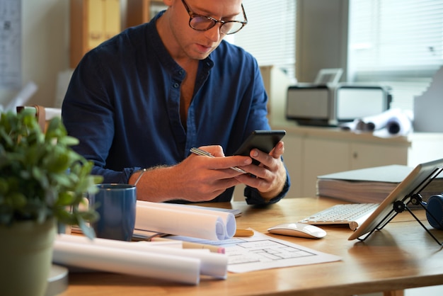 Caucasian man sitting at desk with rolled-up papers and plans and using smartphone