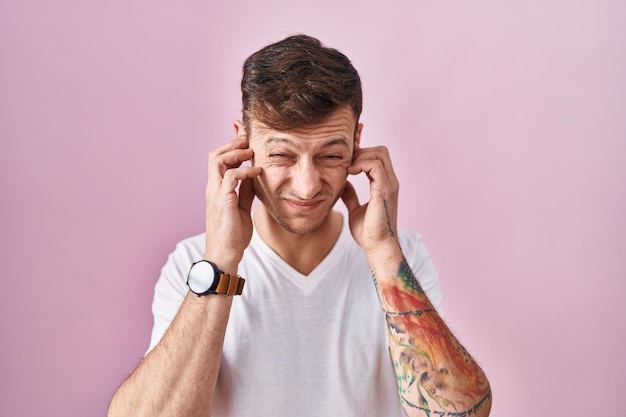 Caucasian man standing over pink background covering ears with fingers with annoyed expression for the noise of loud music. deaf concept.