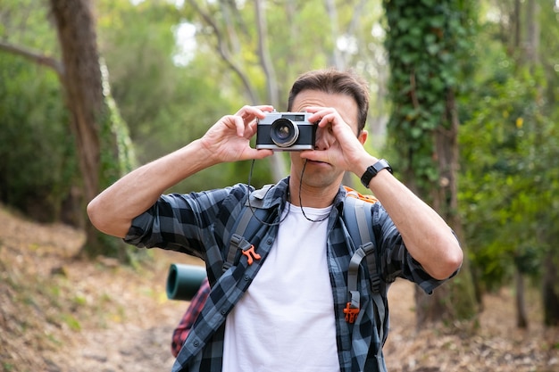 Caucasian man taking photo of nature with camera and standing on forest road. Young male traveler walking or hiking in forest. Tourism, adventure and summer vacation concept