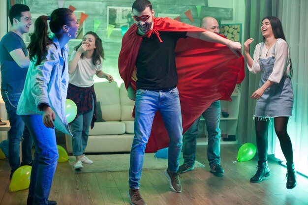 Кавказский человек в костюме супергероя прыгает в воздухе на вечеринке друзей.