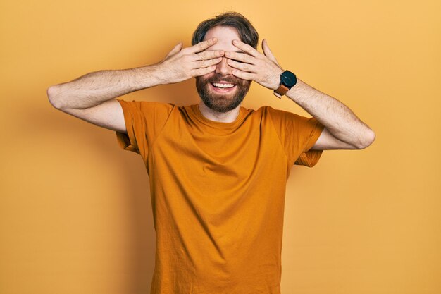 Caucasian man with beard wearing casual yellow t shirt covering eyes with hands smiling cheerful and funny. blind concept.