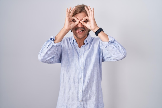 Caucasian man with mustache standing over white background doing ok gesture like binoculars sticking tongue out eyes looking through fingers crazy expression