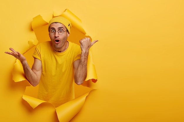 Foto gratuita l'uomo caucasico con l'espressione scioccata punta il pollice a destra su uno spazio vuoto, indossa cappello e maglietta gialli, grandi occhiali rotondi