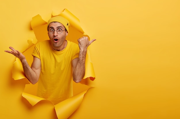 Free photo caucasian man with shocked expression points thumb right on blank space, wears yellow hat and t shirt, big round glasses