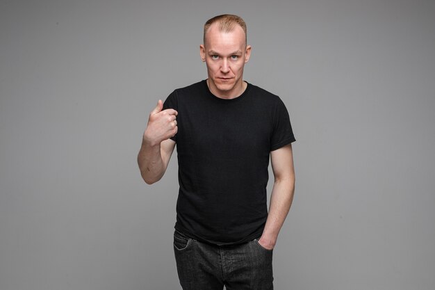 Caucasian man with short fair hair wearing a black t-shirt and jeans points to himself with a finger