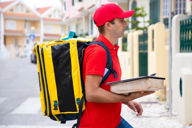 Caucasian postman holding parcel and delivering order. Side view of deliveryman in red cap and shirt going to house of recipient.