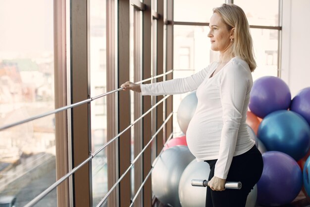 Кавказская беременная женщина в третьем триместре занимается фитнесом в помещении Женщина в яркой фитнес-студии с большими окнами Блондинка в белой рубашке и черных леггинсах