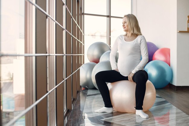 Caucasian pregnant woman in third trimester doing fitness excercise indoors Woman in bright fitness studio with big windows Blonde woman wearing whit shirt and black leggins