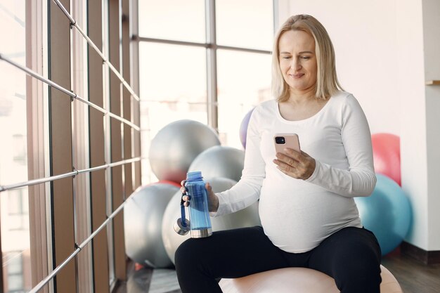 Caucasian pregnant woman in third trimester using a phone a holding a bottle with water Woman in bright fitness studio with big windows Blonde woman wearing whit shirt and black leggins