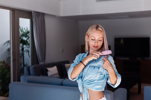 Caucasian pretty blonde short hair woman in denim jacket brushing hair with pink hairbrush at home