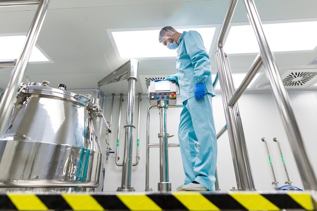 Caucasian scientist in blue lab suit and gloves configure control panel near steel tank look on panel black and yellow stripes