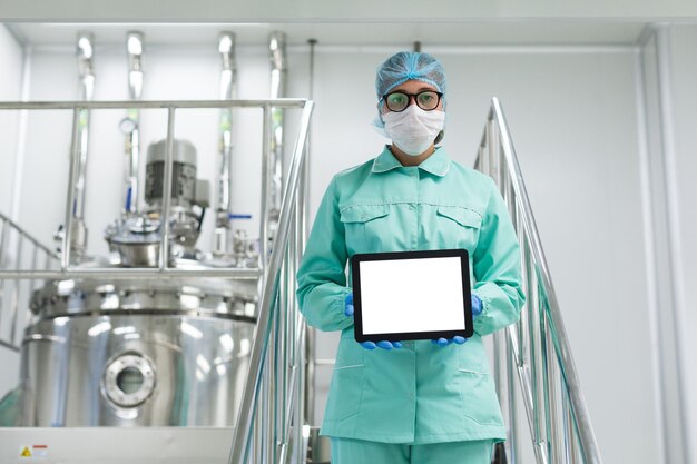 Caucasian scientist in blue lab uniform and glasses stand on chromed stairs and hold empty tablet towards camera