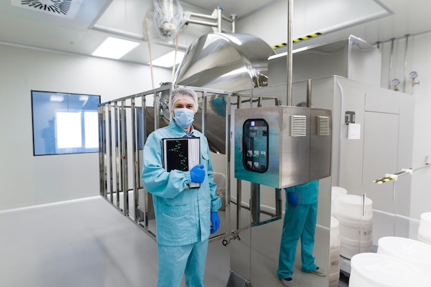 Caucasian scientist in blue lab uniform stand near big steel machine with control panel look at camera