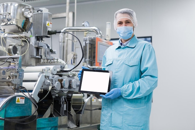 Caucasian scientist in blue lab uniform stand near manufacture machine with shafts show empty tablet