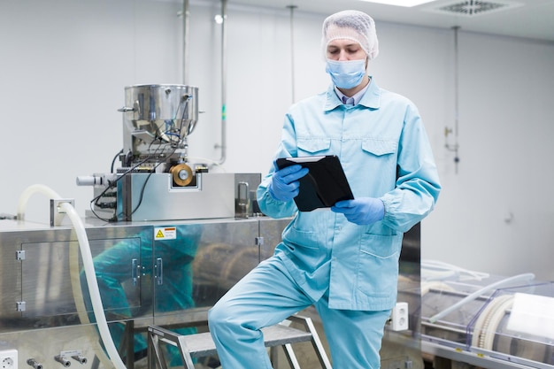 파란색 실험복을 입은 백인 과학자 남자는 클린룸에 있는 큰 금속 기계와 함께 카메라를 쳐다보는 크롬 사다리 위에 서 있습니다.