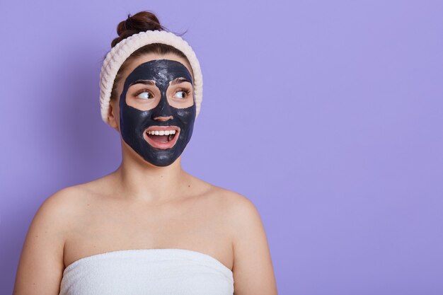 Caucasian surprised beautiful woman with towel on her body with black cleansing mask on face isolated over lilac wall, looking away with widely opened mouth, copy space