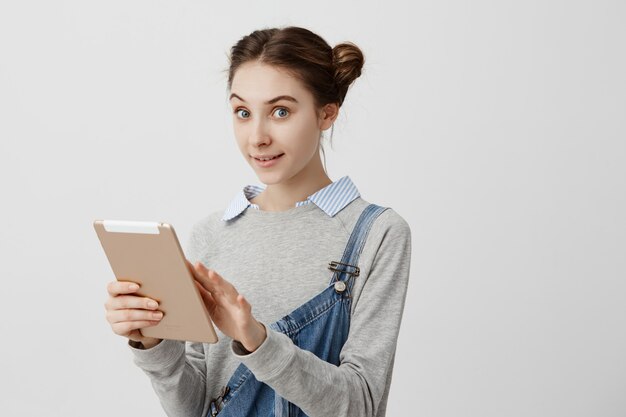 Caucasian woman being girlish with odango buns looking  with delighted gaze holding notebook. Positive emotions of female buyer testing new digital tablet. Technology, future