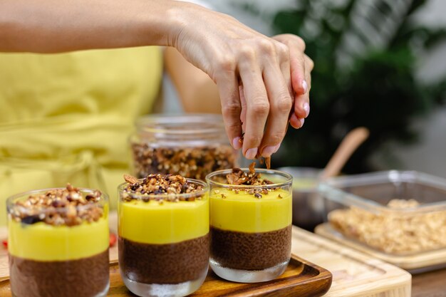 Caucasian woman in kitchen makes chia puddings with mango jam. Desert made of almond milk, chia seeds, cocoa, mango jam and granola.