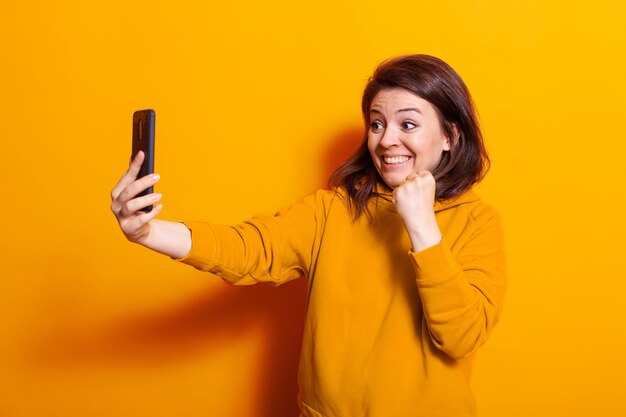 Caucasian woman looking at smartphone and smiling in studio. Joyful person holding mobile phone and watching screen while clenching fist and feeling happy on camera. Cheerful adult