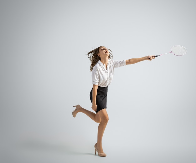Caucasian woman in office clothes plays badminton on grey