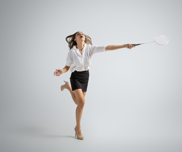 Caucasian woman in office clothes plays badminton isolated on grey wall